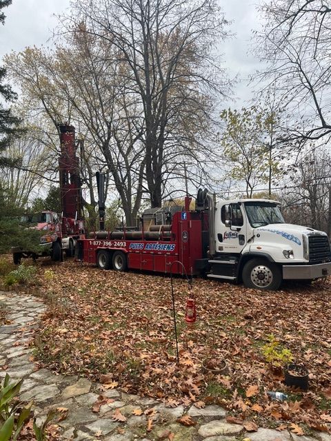 Camion de forage de puits sur une propriété résidentielle, rouge et blanc, feuilles au sol.