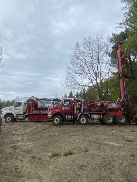 Deux camions de forage stationnés sur un terrain vague sous un ciel nuageux. Un rouge, un blanc.