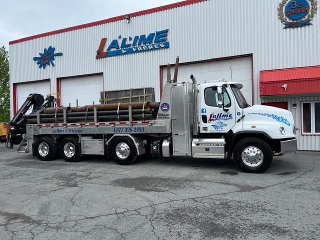 Camion blanc L'Alime Outillage avec une grue chargée de tuyaux, stationné devant un bâtiment portant le logo de l'entreprise.