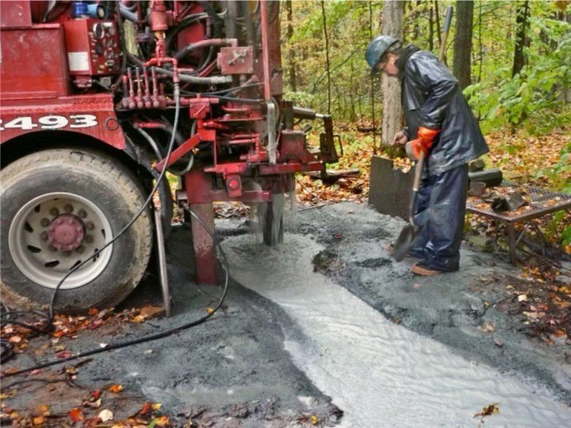 Un homme en ciré pelle de la boue près d'une plateforme de forage dans une zone boisée.