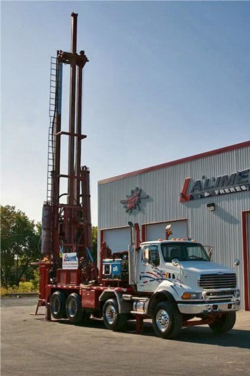 Une foreuse montée sur un camion blanc devant un bâtiment rouge et blanc par une journée ensoleillée.