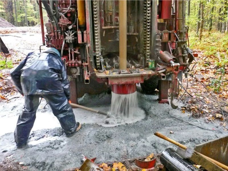 Personne en tenue de pluie travaillant sur une plateforme de forage, pulvérisant de l'eau et des débris. Environnement forestier extérieur.