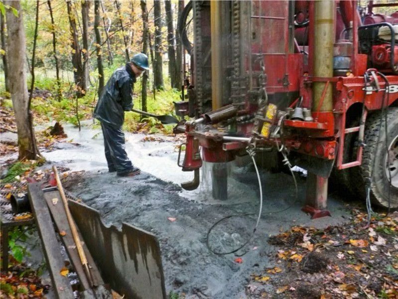 Une personne utilise du matériel de forage dans un environnement forestier boueux ; machines rouges, boue grise.