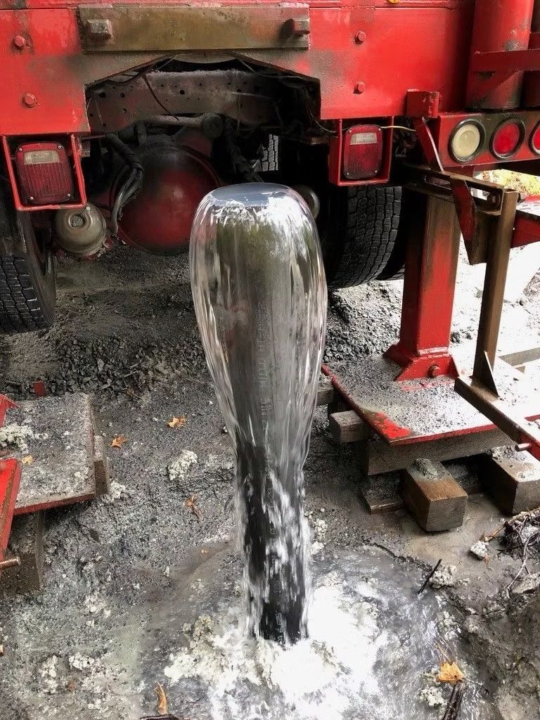 De l'eau jaillit d'un tuyau de puits, avec un camion rouge en arrière-plan, entouré de terre et d'outils.