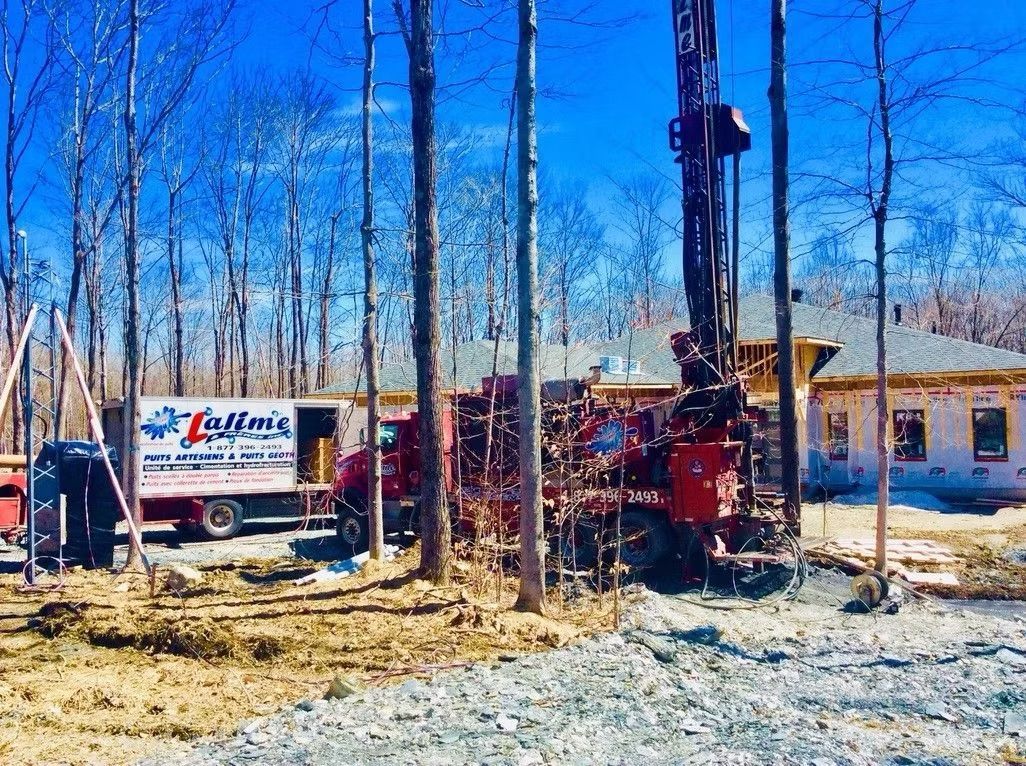 Une foreuse rouge et un camion blanc de la compagnie « Lufthens » se trouvent dans une zone boisée où une maison est en construction.