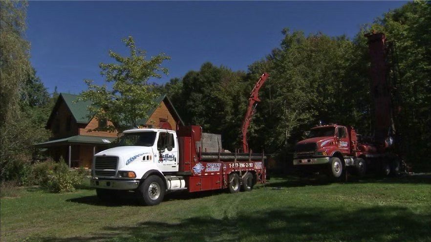 Deux gros camions, l'un blanc, l'autre rouge, chargés de matériel, sont stationnés dans une cour herbeuse, près d'une maison en bois et d'arbres, par une journée ensoleillée.
