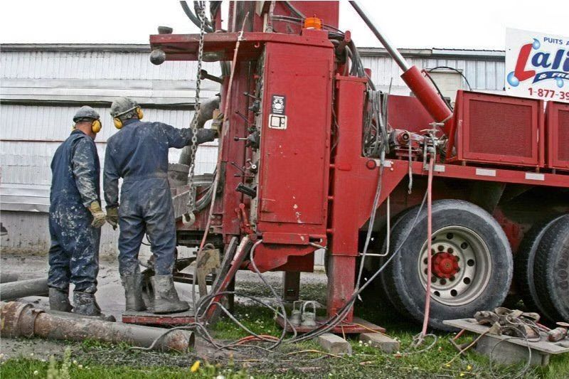 Deux ouvriers manœuvrant une foreuse rouge en extérieur. L'un d'eux se trouve près d'une canalisation ; tous deux portent des combinaisons et des casques de chantier.