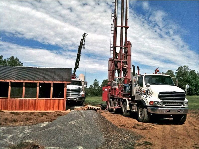 Deux gros camions sur un champ herbeux, l'un d'eux transportant une foreuse, à côté d'un petit bâtiment en bois et d'un tas de gravier.