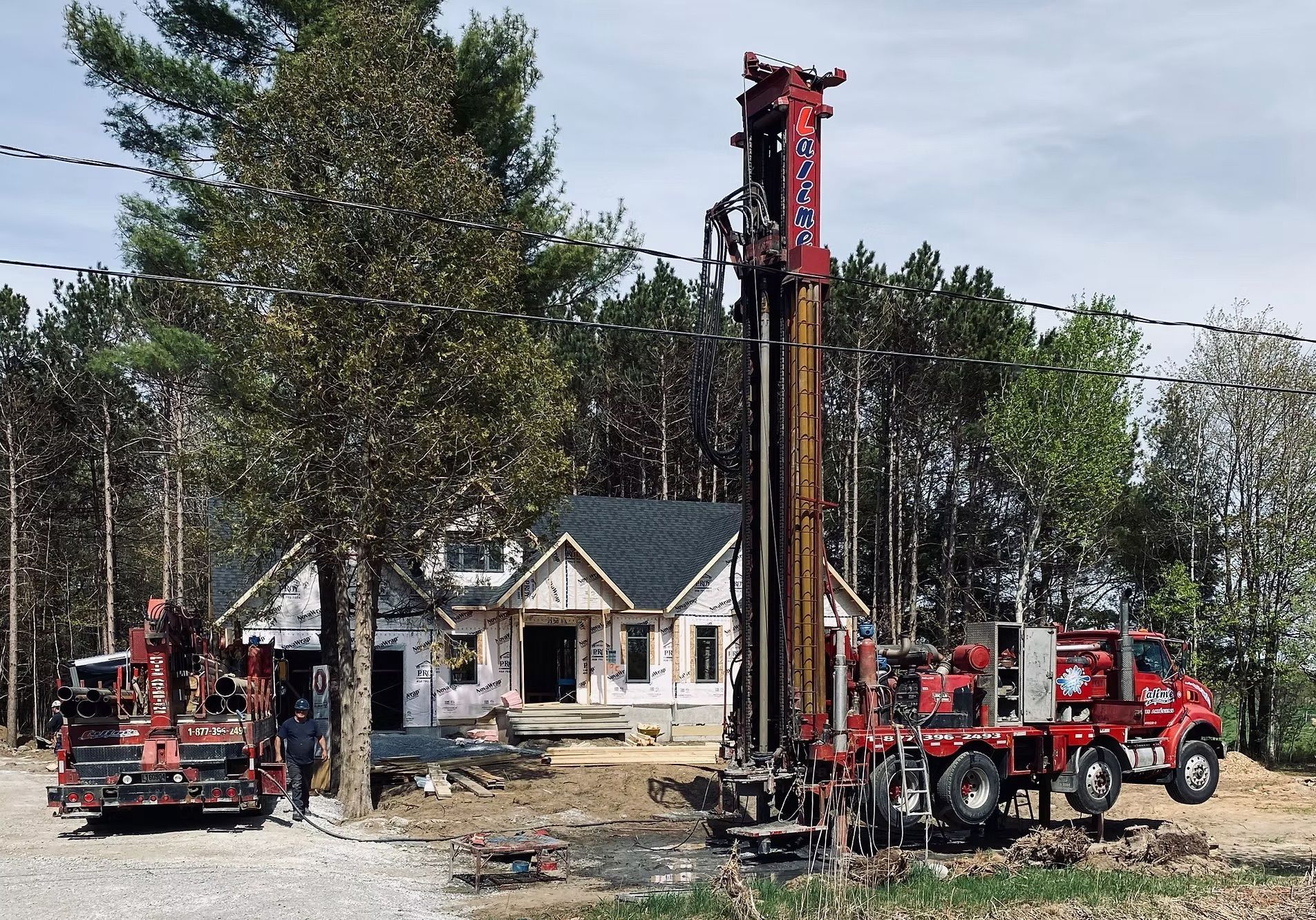 Deux camions de forage rouges près d'une maison en construction. Arbres en arrière-plan, journée ensoleillée.