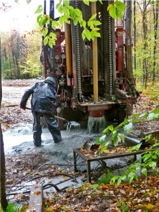 Un homme manœuvre une foreuse dans une zone boisée, de l'eau jaillit.