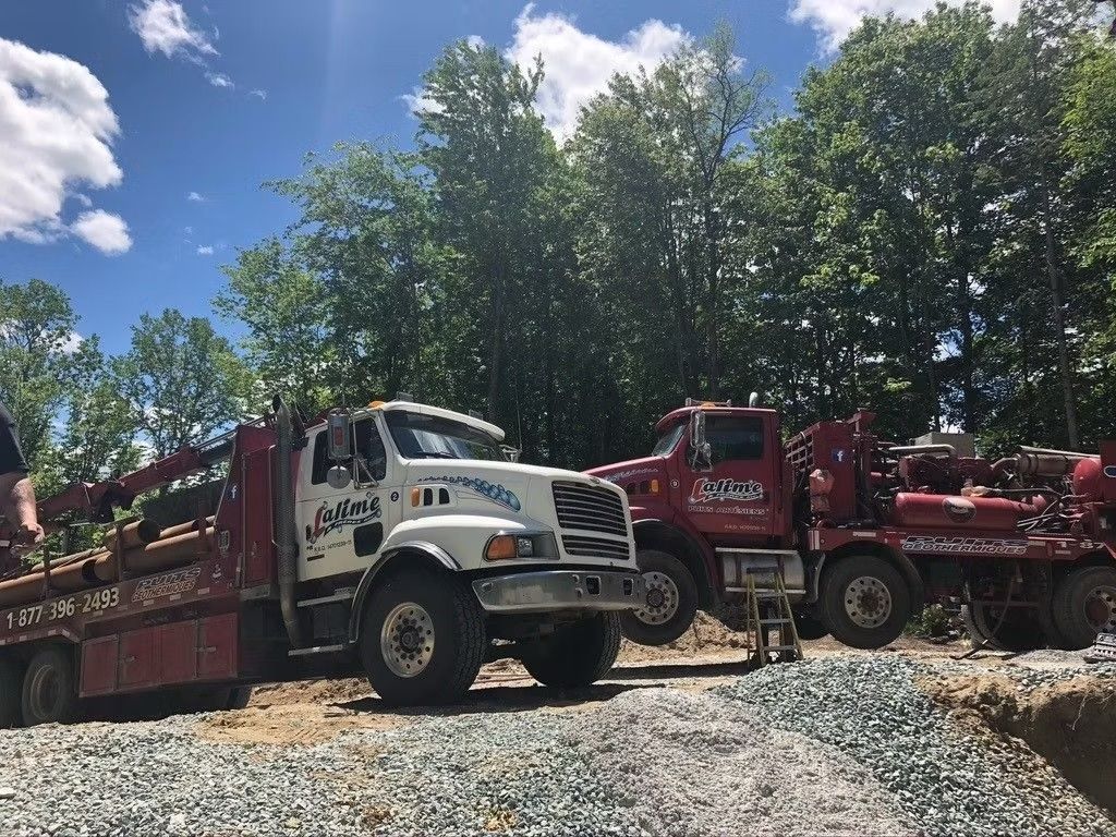 Deux camions de chantier sur du gravier, un blanc et un rouge, dans une zone boisée sous un ciel bleu.