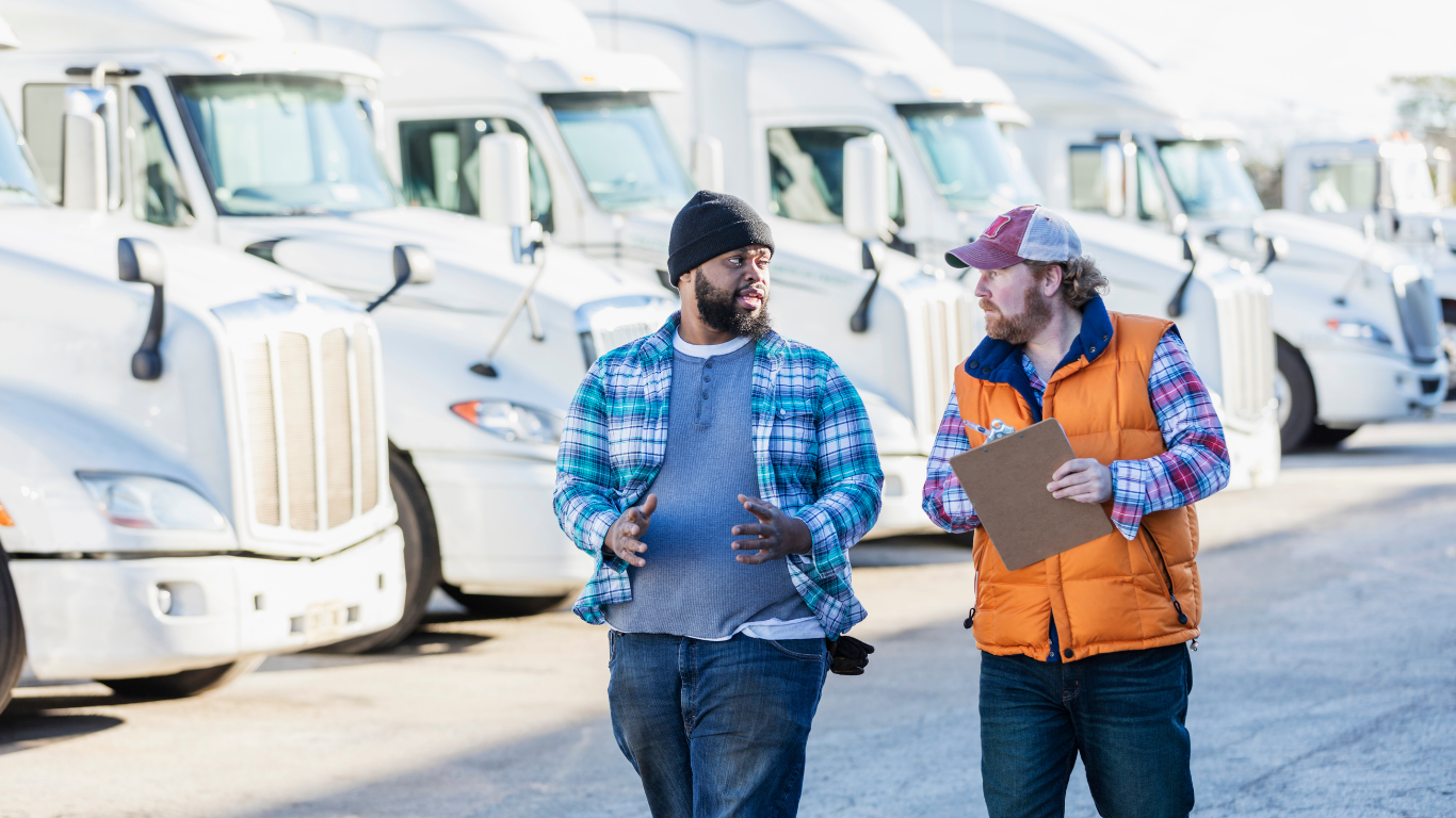 Two men talking in front of a line of parked white semi-trucks. One holds a clipboard.