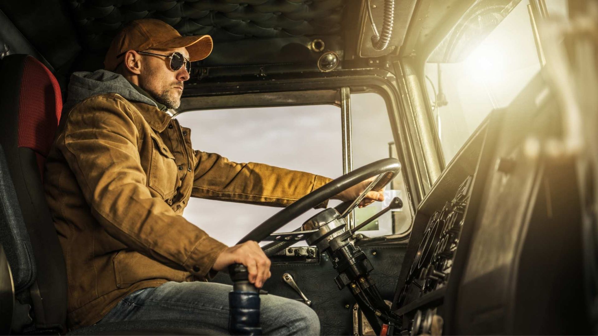Man driving a semi-truck; sunny, daytime; brown jacket, cap, and sunglasses.