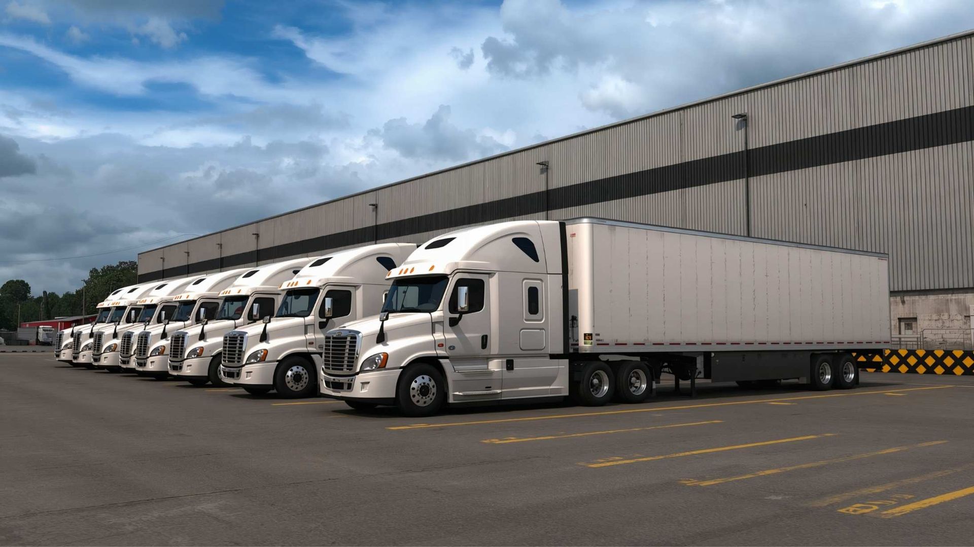 Row of white semi-trucks parked in front of a large warehouse on a cloudy day.