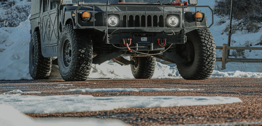 A dark Humvee parked on a gravel surface partially covered in snow against a snowy, mountainous background.