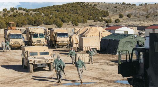Soldiers in camouflage uniforms move through a sandy military encampment featuring tan trucks and a large green tent.