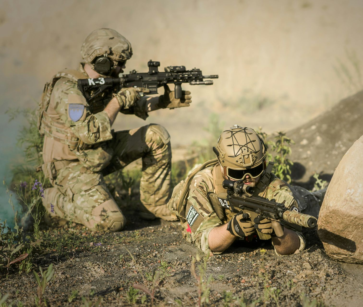 Two soldiers in camouflage gear aim weapons near a rock in a dusty outdoor setting.