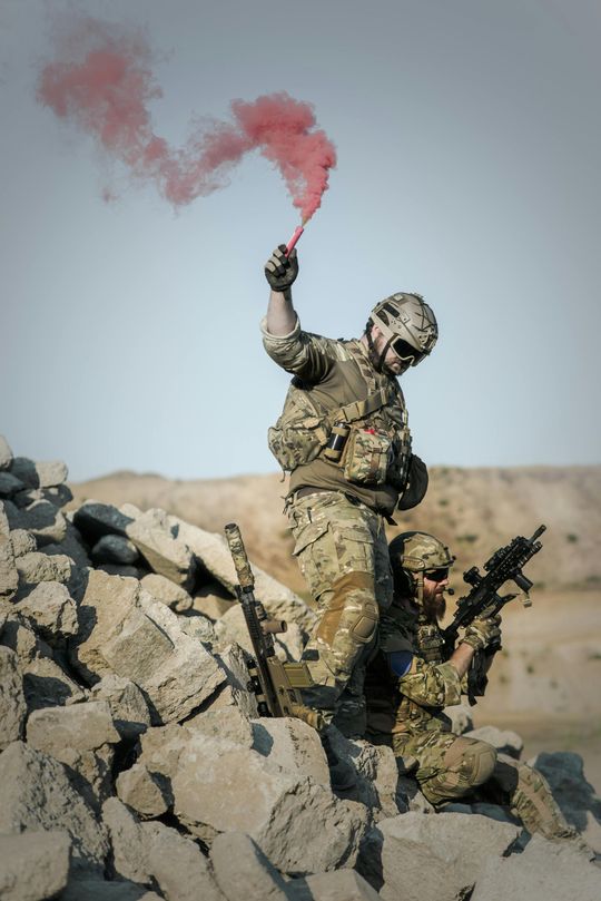 Two soldiers in camouflage gear crouch on a rocky mound, one holding a smoking red flare aloft against a pale sky.