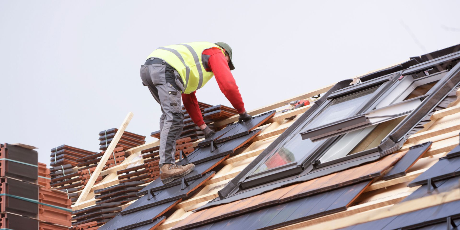 Roofer in yellow vest lays tiles on a rooftop near skylights.