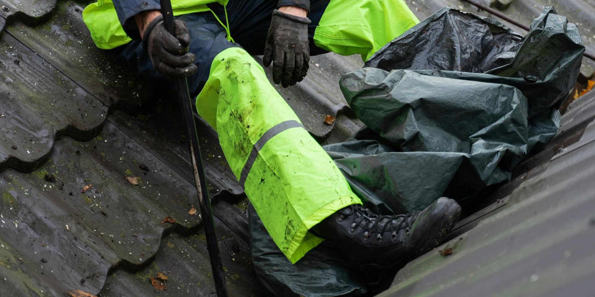 Gloved hand cleaning autumn leaves from a roof gutter on a house.