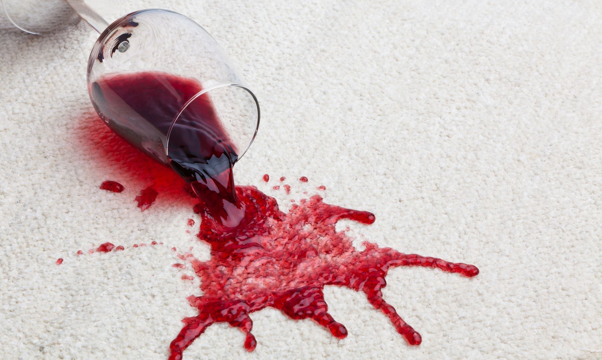 A glass of red wine has spilled on a white carpet.