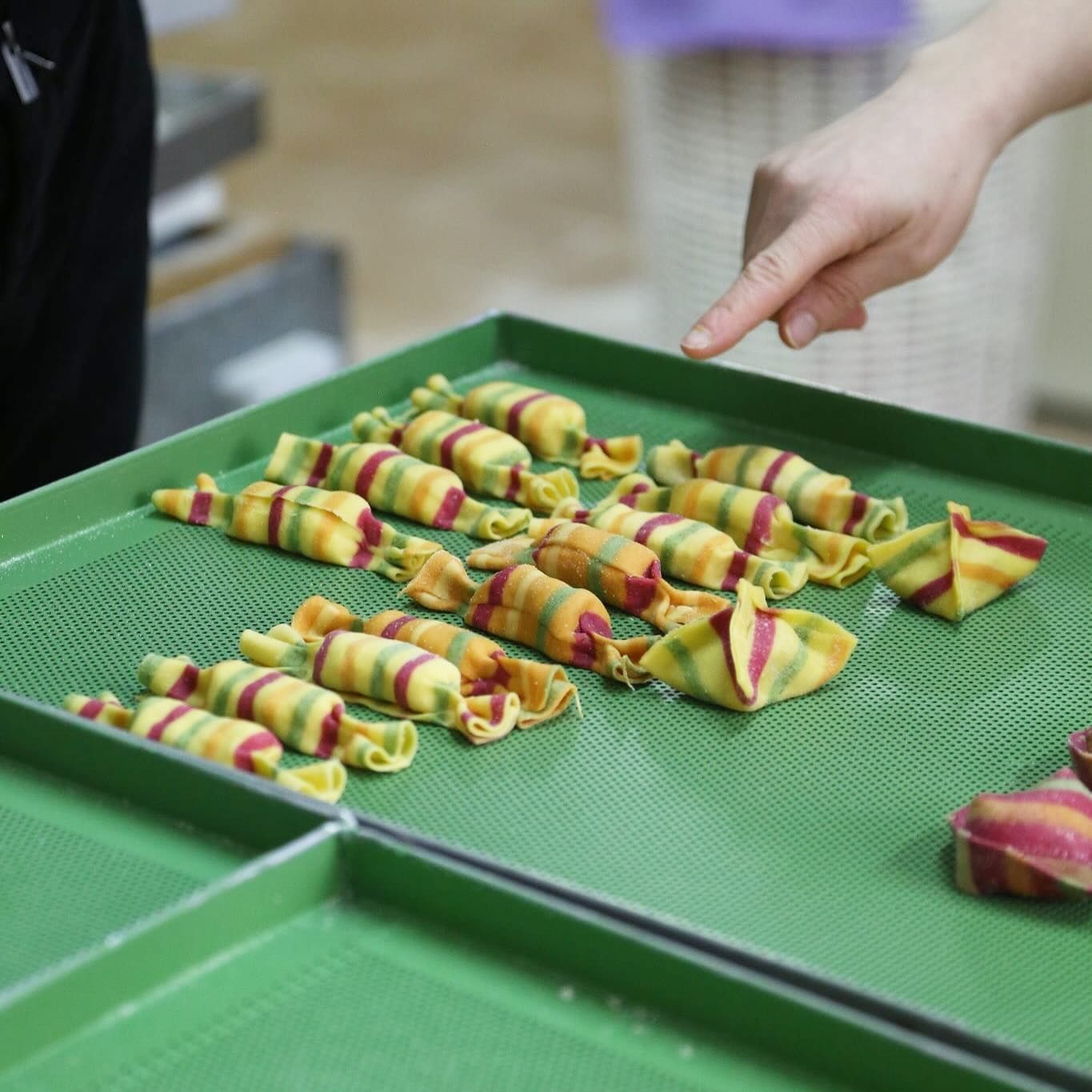 Mano che indica un vassoio di pasta colorata a forma di caramella, appoggiata su una superficie verde.