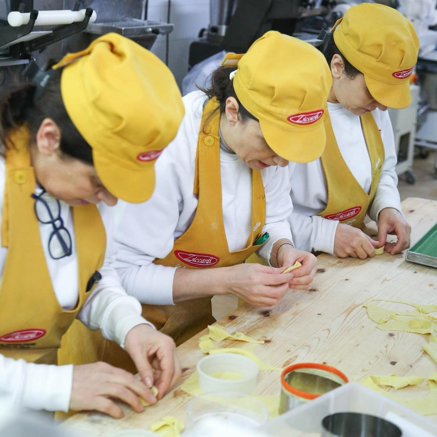 Tre persone con cappelli e grembiuli gialli preparano la pasta su un tavolo di legno.