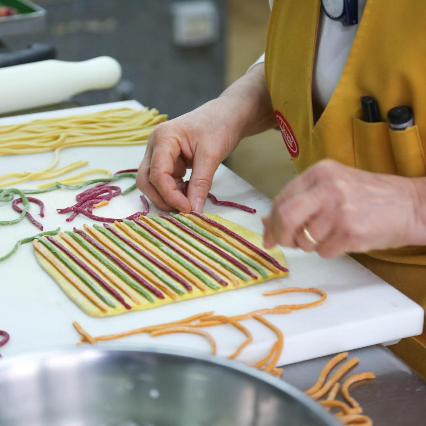 Persona che dispone fili di pasta colorata su una superficie bianca, indossando un grembiule giallo.