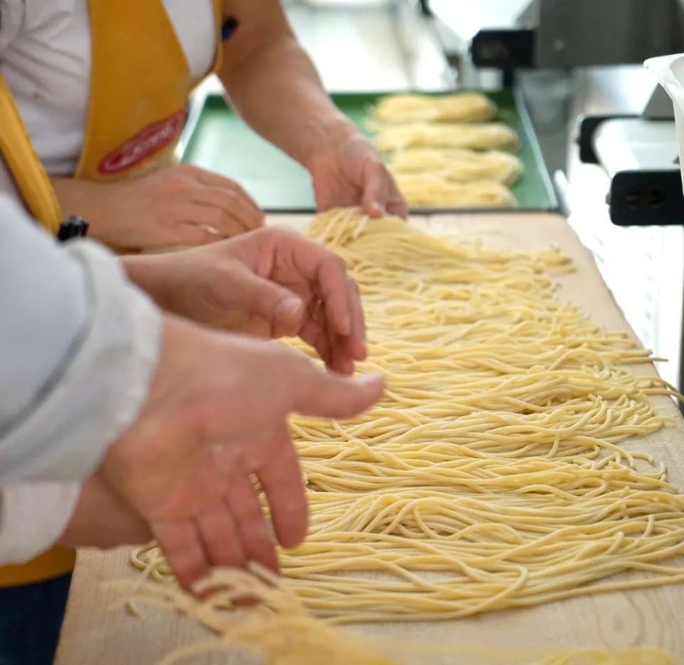 Mani che dispongono spaghetti freschi e crudi su una superficie di legno. Sullo sfondo, un altro vassoio di pasta.