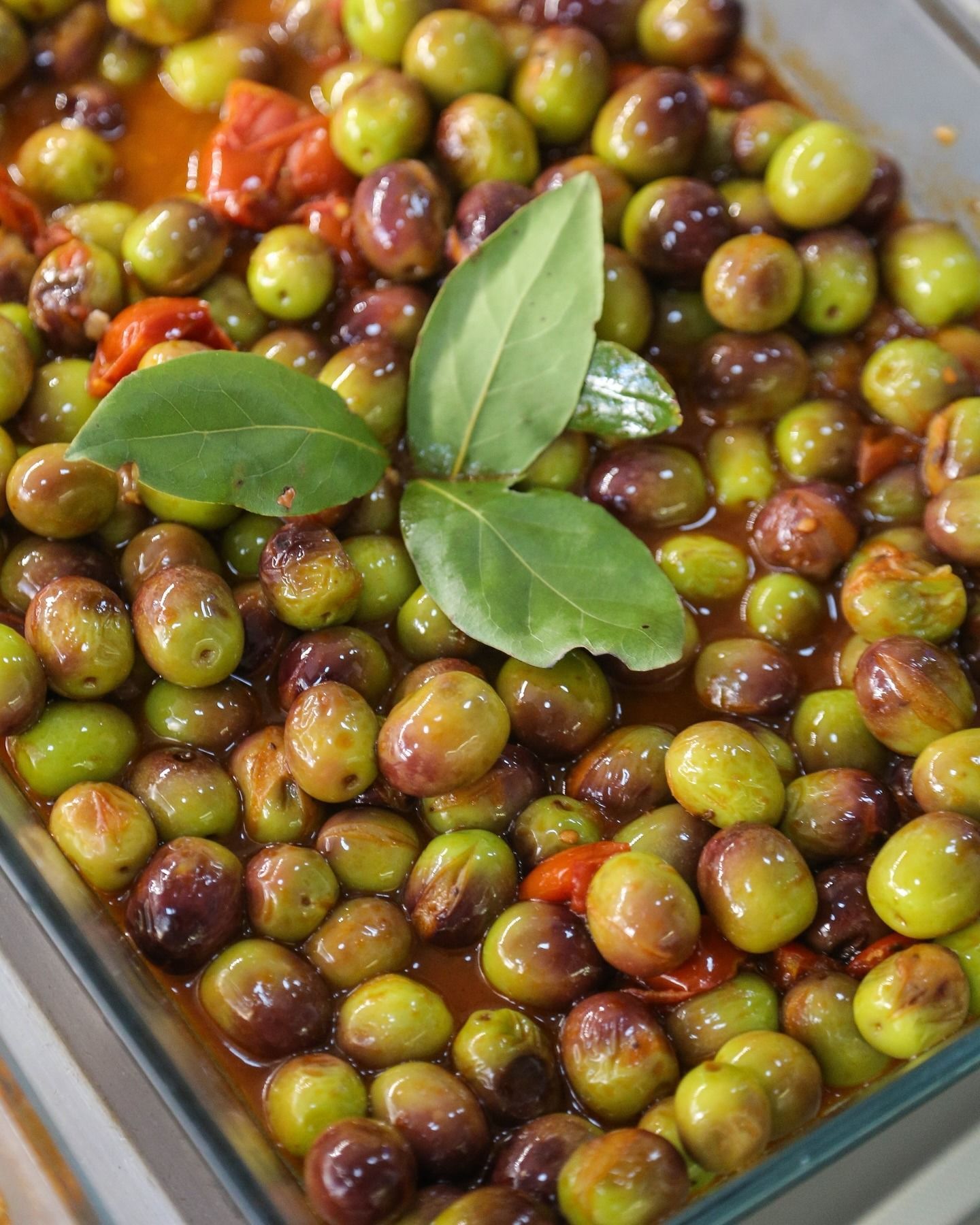 Piatto di vetro con olive verdi e viola in salsa rossa, guarnito con foglie di alloro.