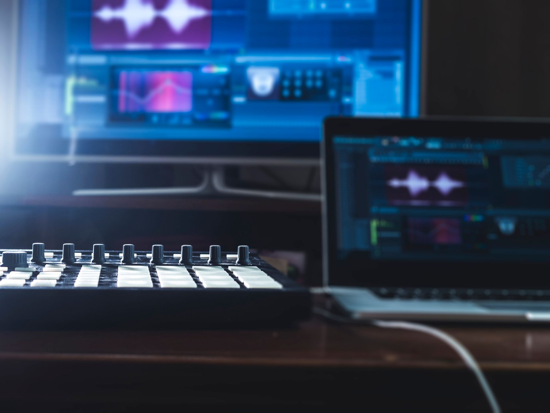 A music production setup featuring a MIDI controller, a laptop, and a monitor displaying audio editing software.