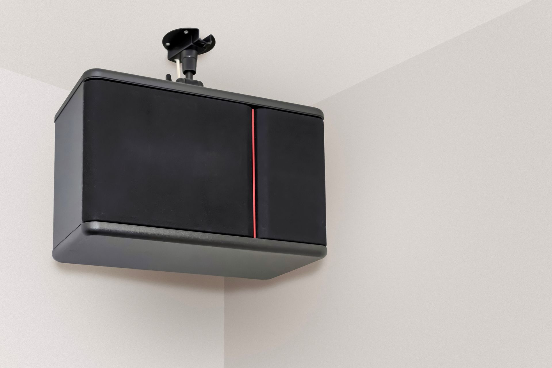 A black, rectangular ceiling-mounted speaker with a red vertical accent line, installed in a white corner.