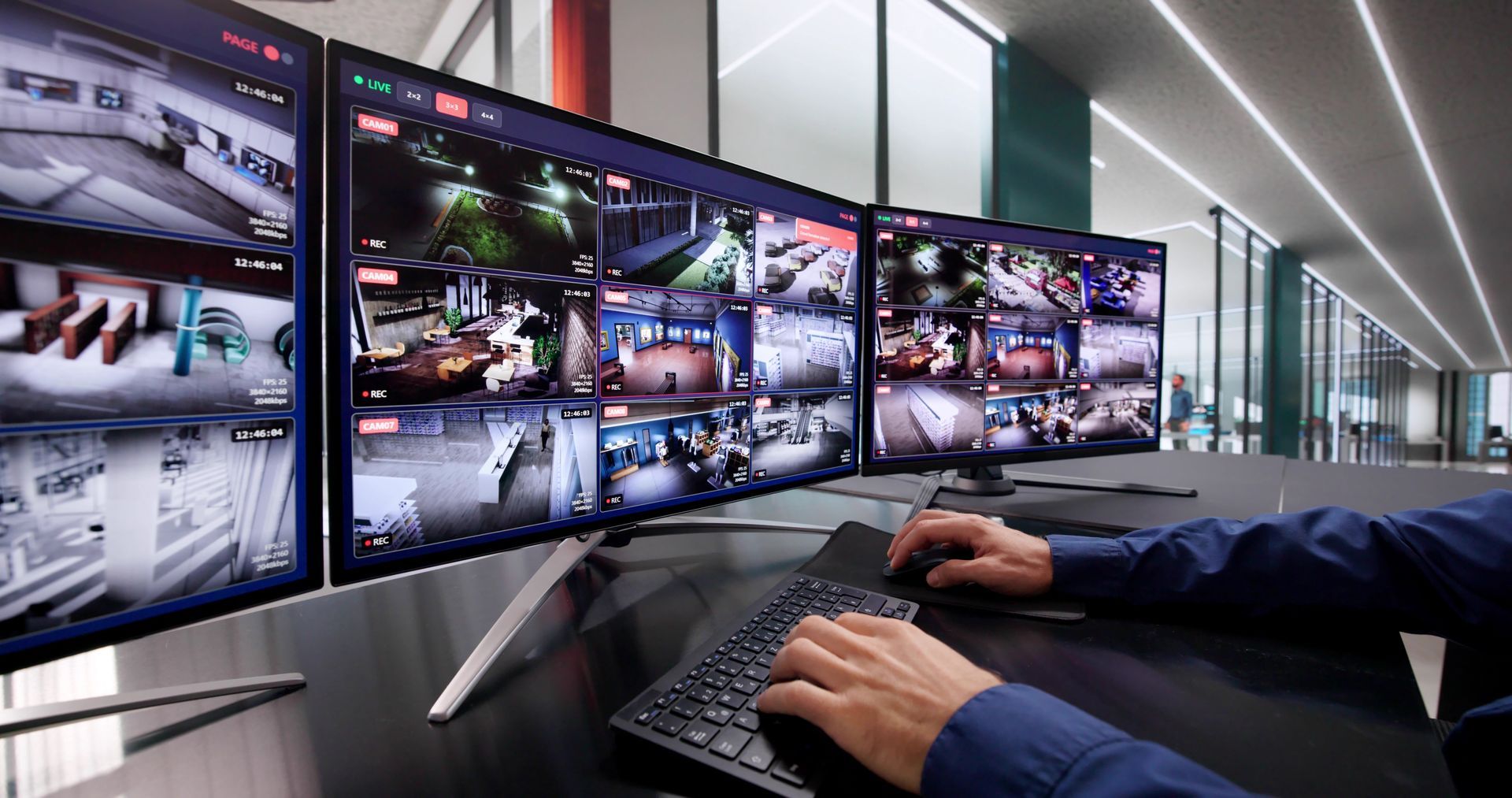 A person uses a keyboard to monitor multiple security camera feeds displayed on a bank of three computer screens.