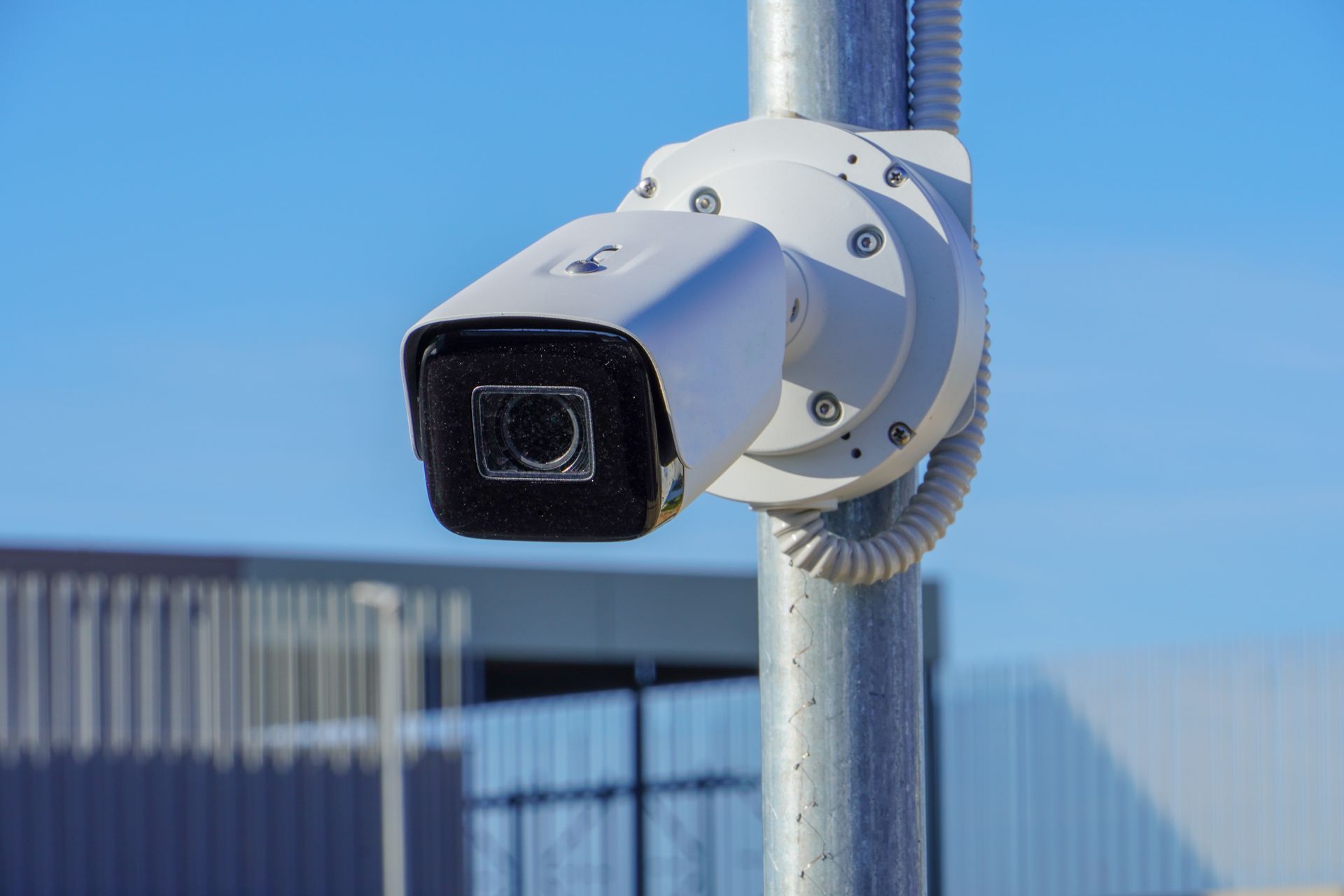 A white security camera mounted on a metal pole against a blue sky.