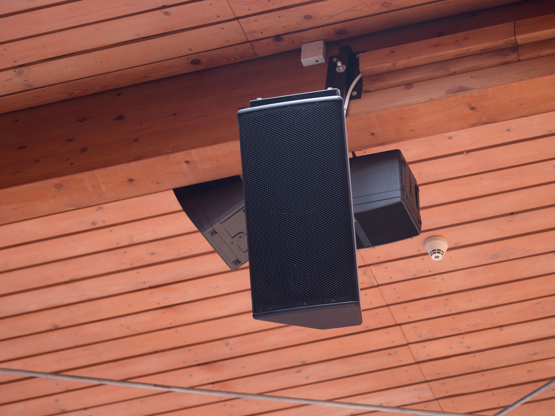 Two black speakers and a small smoke detector are mounted to a slatted wooden ceiling.