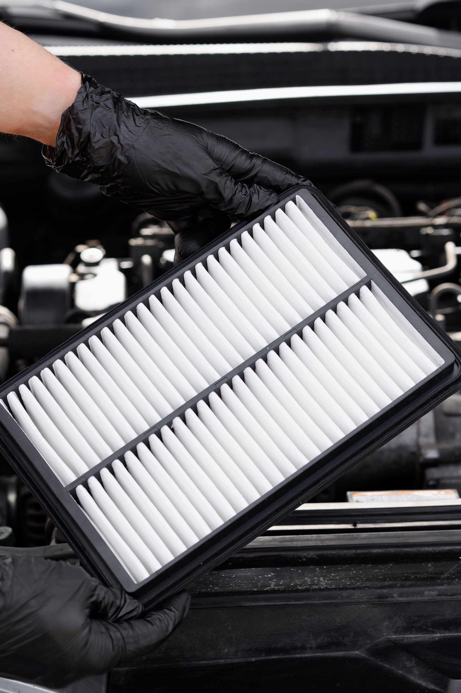 Hand holding a new rectangular air filter for a car, next to the open engine compartment.
