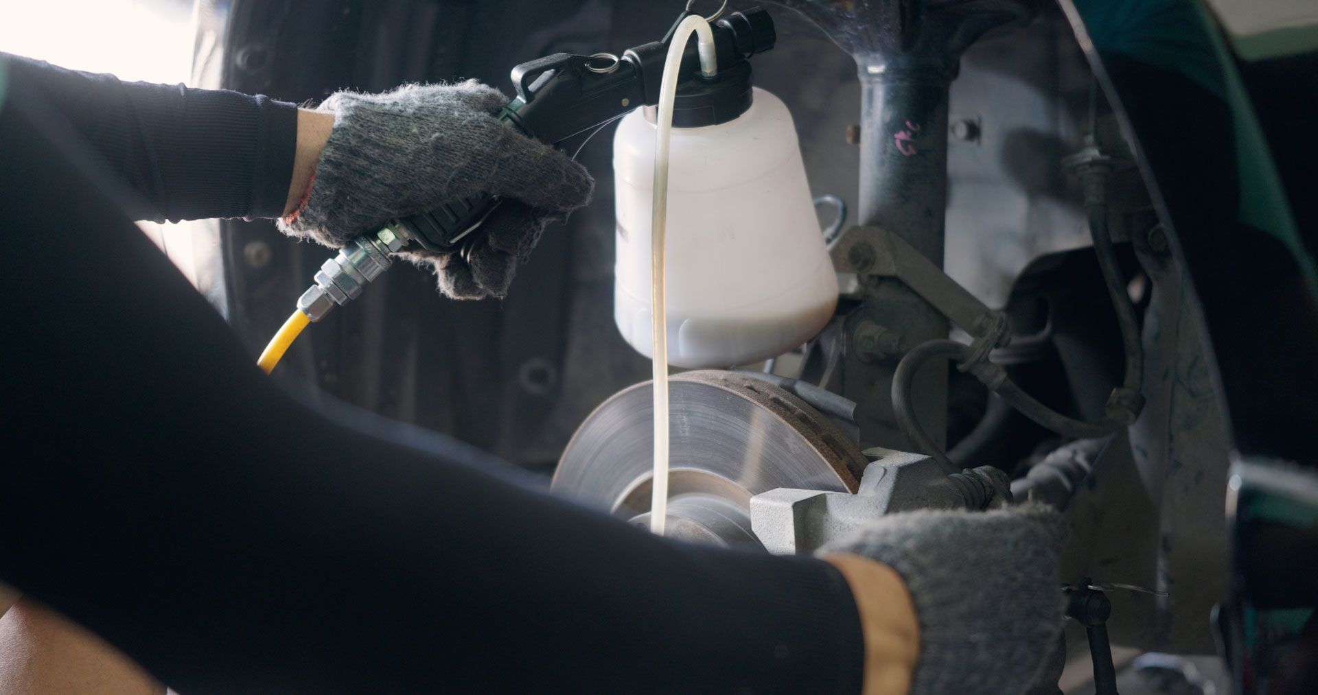 Person bleeding brakes on a vehicle with a fluid reservoir, wearing gloves in a garage.
