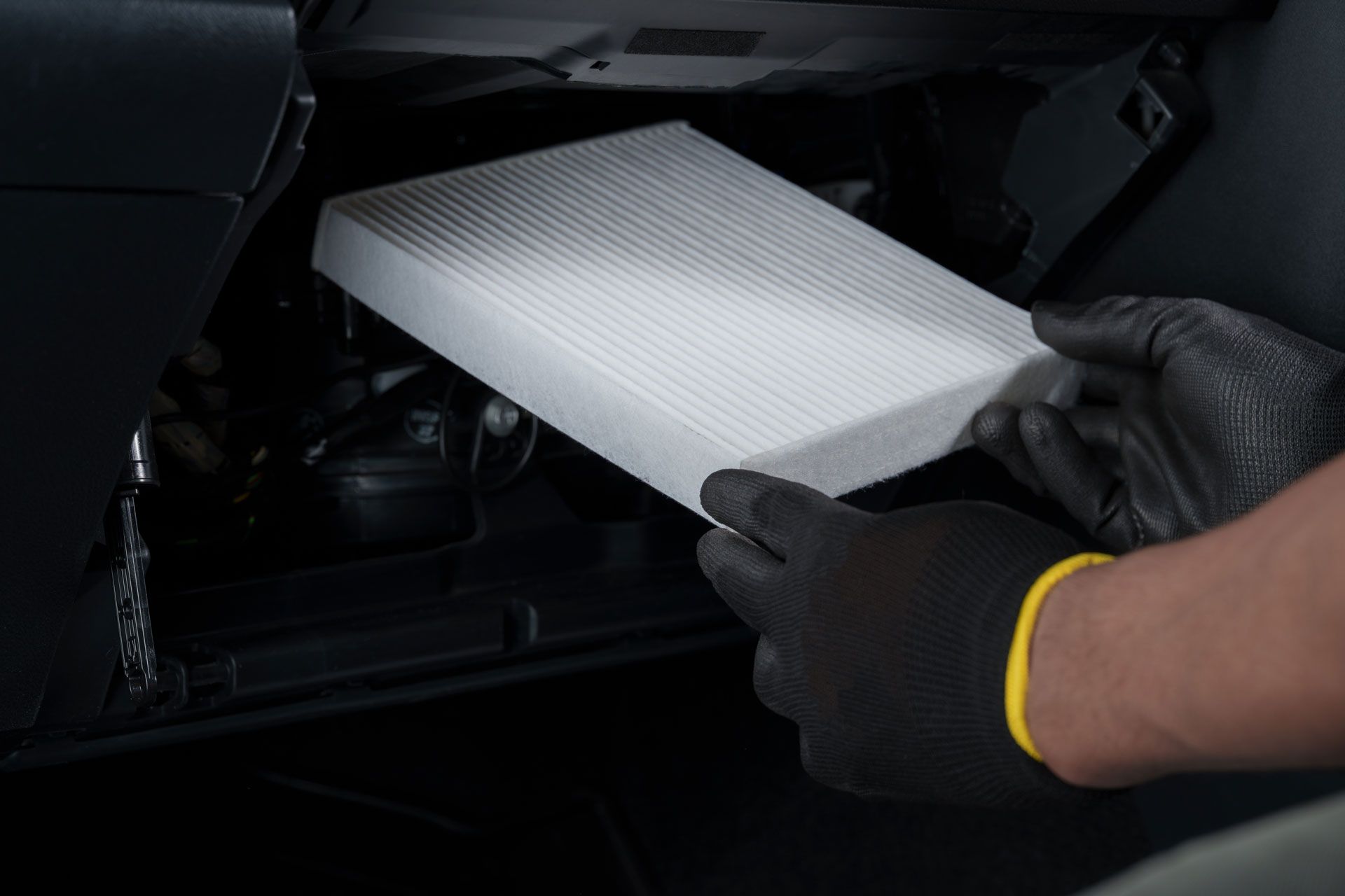 Gloved hands holding a new rectangular cabin air filter near its compartment in a car's dashboard.