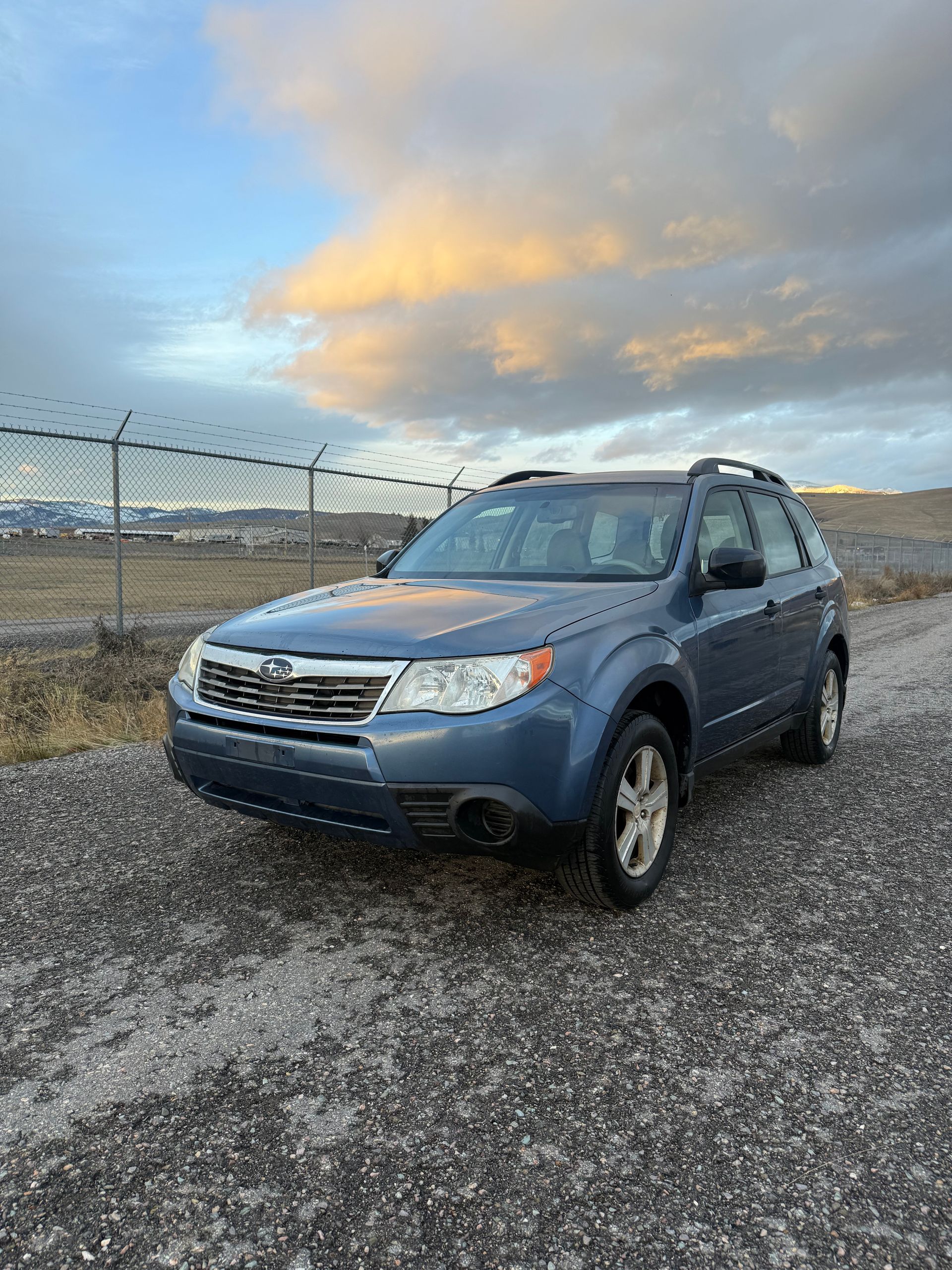 Blue Subaru Forester SUV parked on a gravel road next to a chain-link fence, under a cloudy sky.
