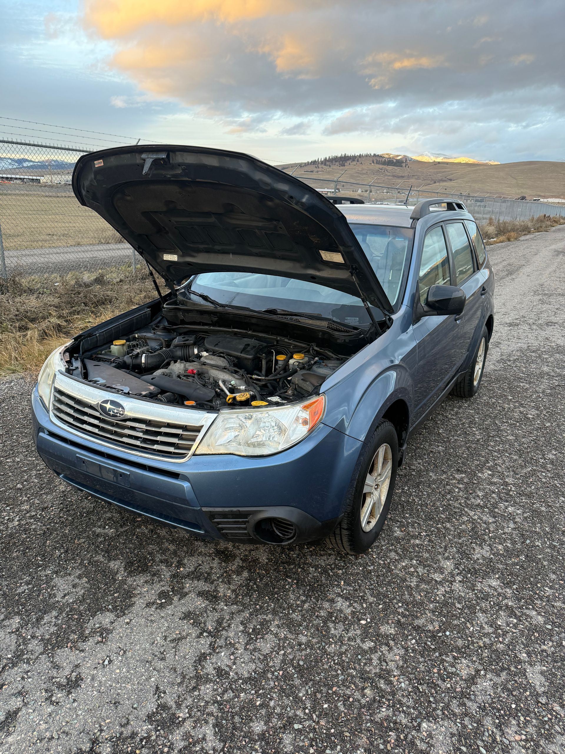Blue Subaru Forester with the hood open on a gravel road, overcast sky.
