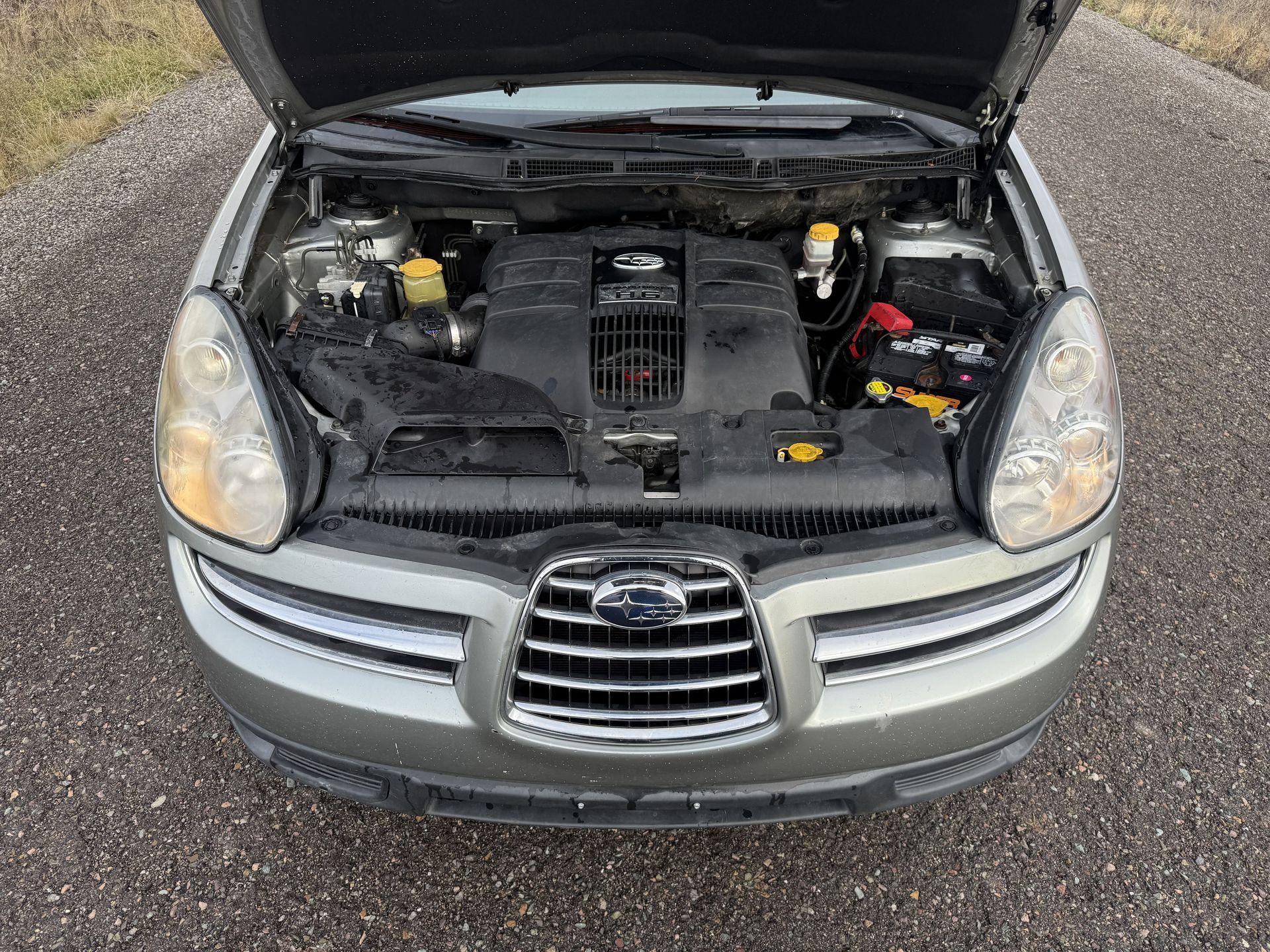 Gray Subaru Tribeca with open hood showing engine bay, parked on pavement.