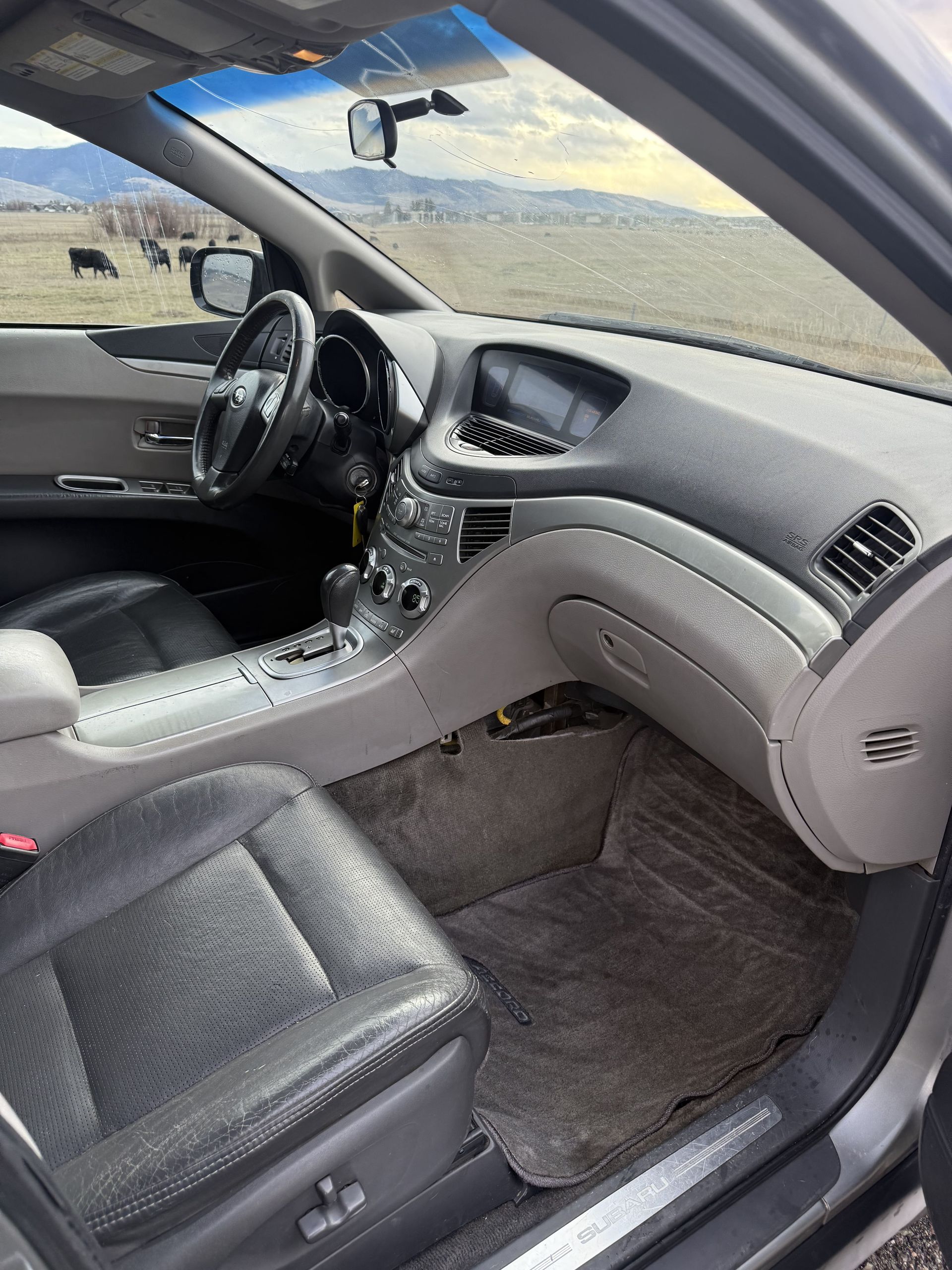 Interior of a gray Subaru Tribeca with black leather seats, dashboard, and a view of a field and mountains.