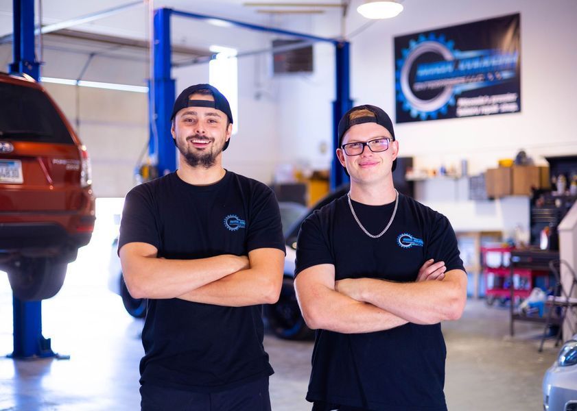 Two men are standing next to each other in a garage with their arms crossed.