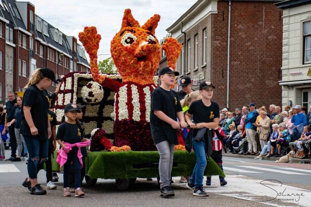 Een groep mensen staat rond een praalwagen van bloemen in een optocht.