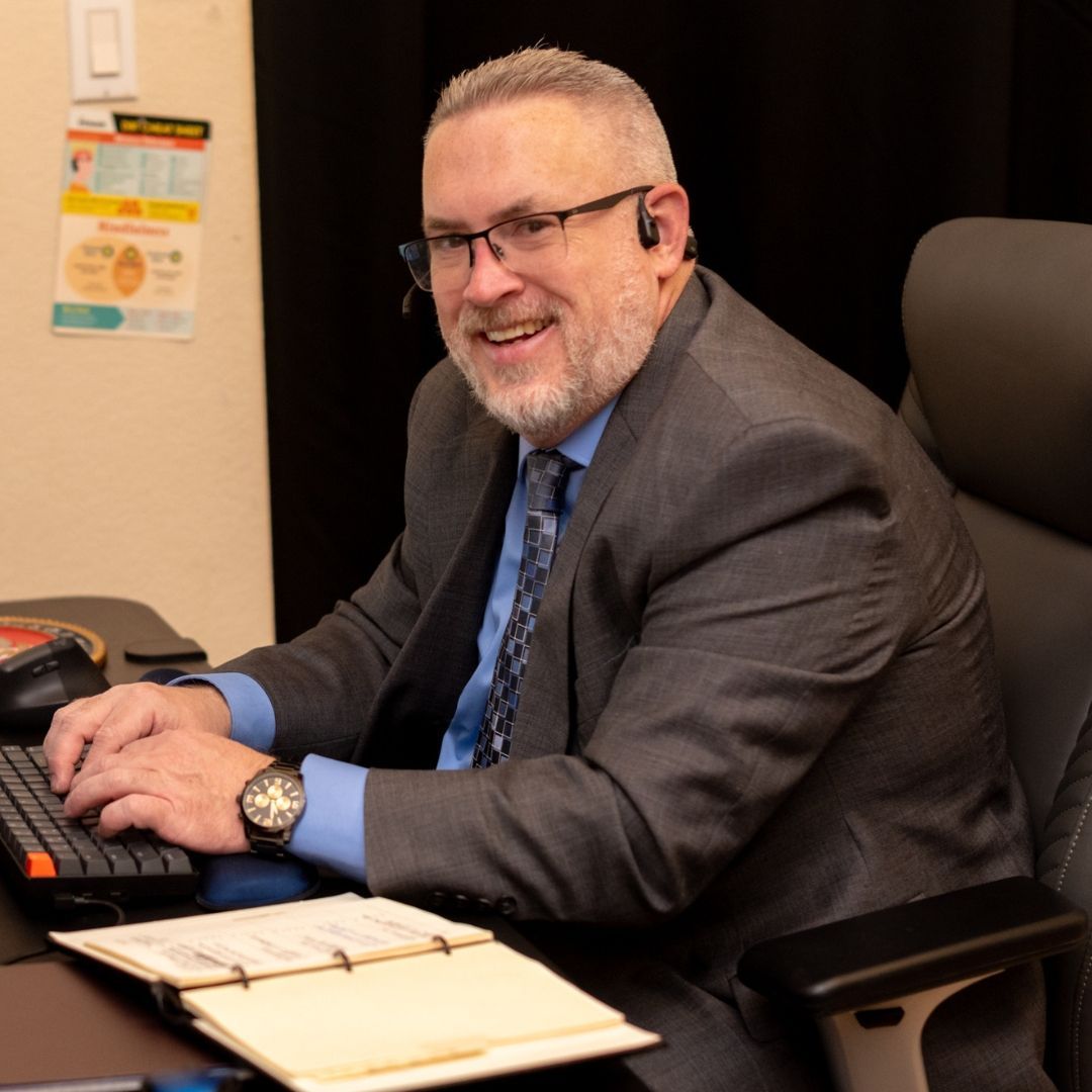 A smiling man in a suit and glasses typing at a desk with an open notebook in an office setting.