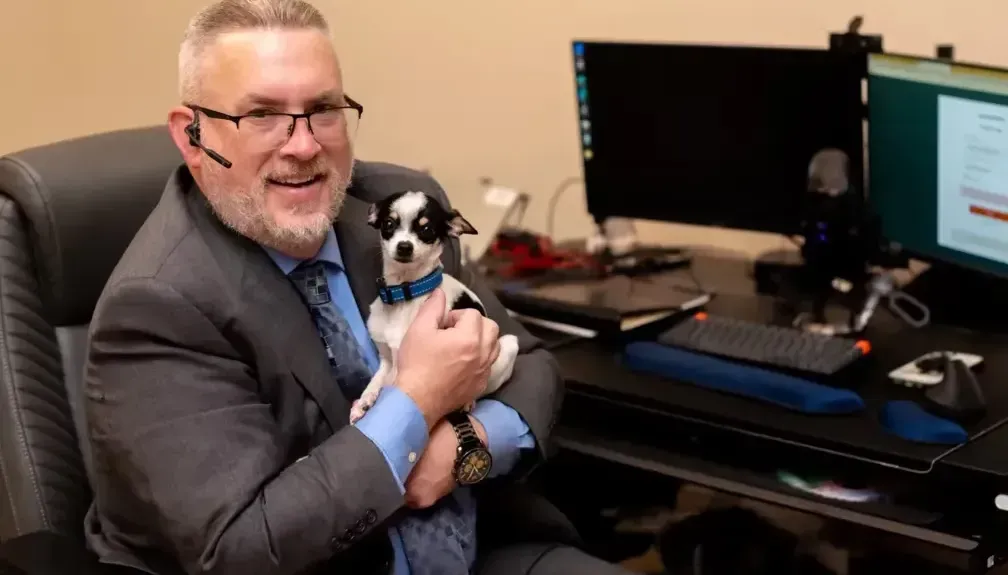 A person in a suit and glasses sits at a desk with two computer monitors, holding a small black and white dog.