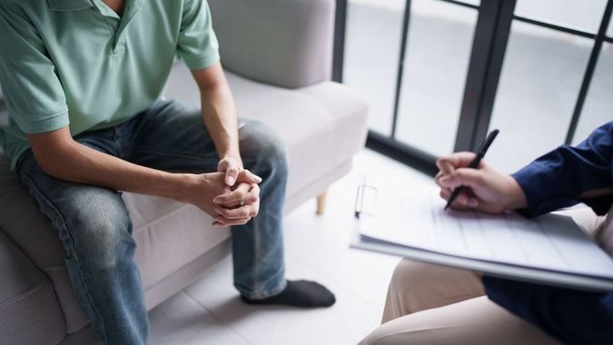 A person in a green polo shirt sits on a sofa while a professional takes notes on a clipboard during a consultation.