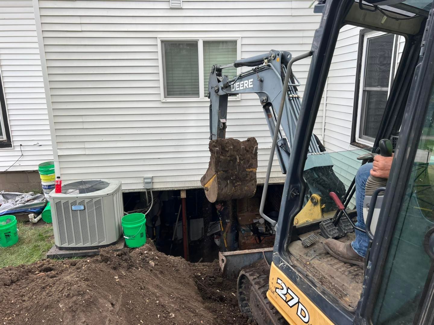 An excavator digging near a house, creating an opening under the structure. A man operates the machine.