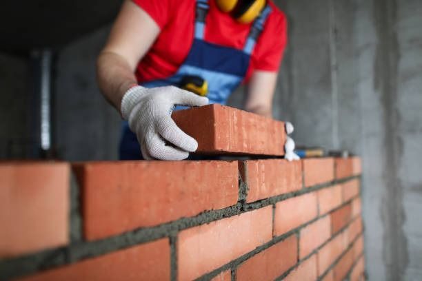 A construction worker in a red shirt and blue overalls places a brick on a partially built brick wall.