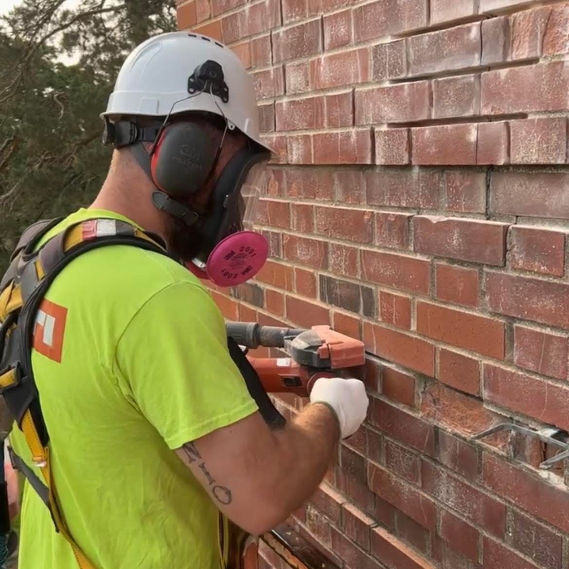 Construction worker grinding brick mortar on a wall; he wears a mask, hard hat, and safety harness.